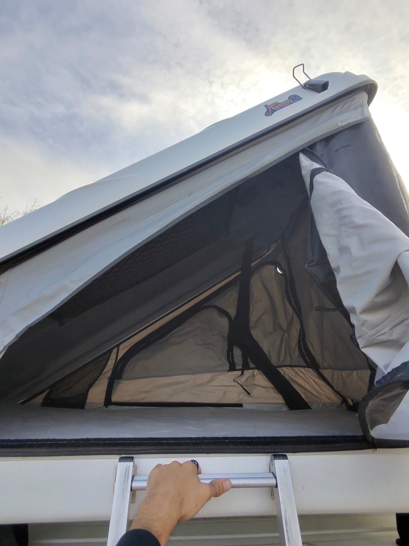Looking up at an open James Baroud rooftop tent on a vehicle, with a hand gripping the ladder. Ideal for an rv rental.