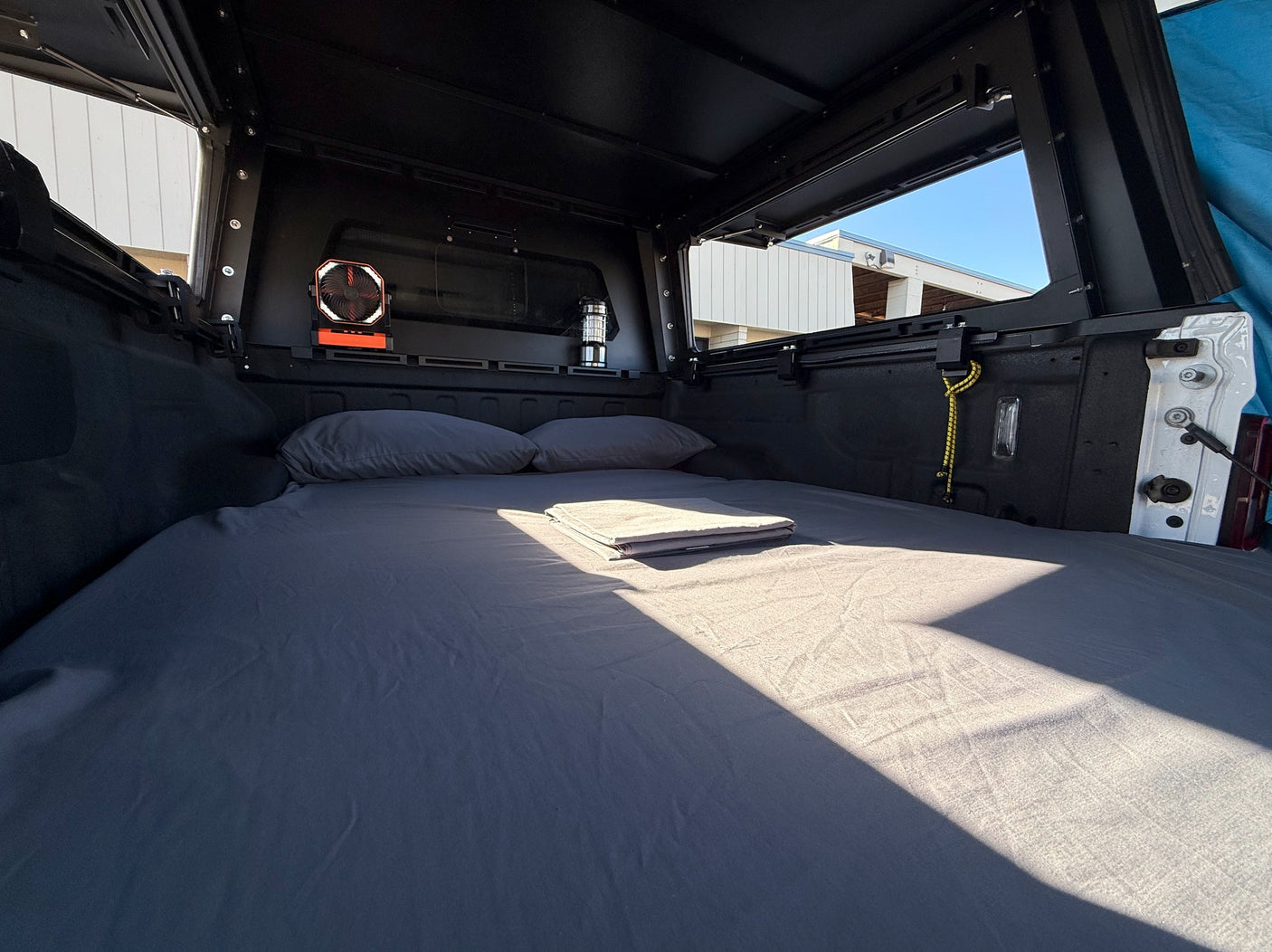 Cozy truck bed camper setup with mattress, pillows, fan & lantern, illuminated by sun. Ideal for overlanding or an RV rental trip.