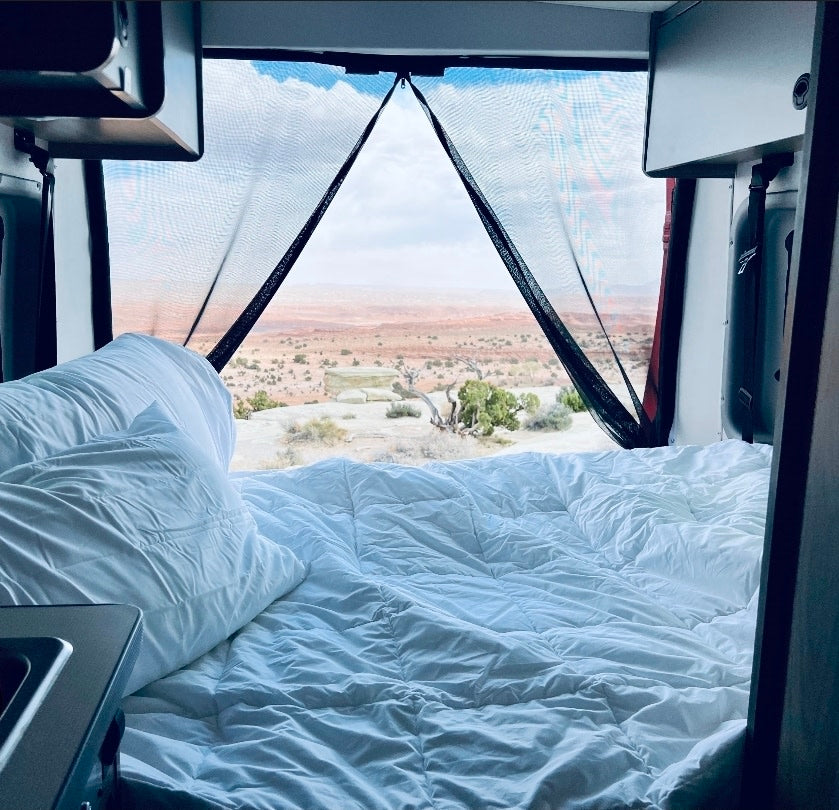 Inside a comfy rv rental, a bed with white linens faces a stunning desert landscape through a mesh window screen, perfect for van life.