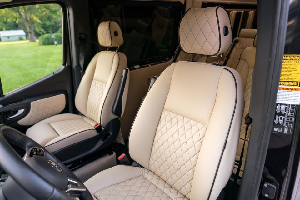 parked on a gravel path with green grass and trees in the background.|Luxury Mercedes-Benz RV rental interior: spacious driver's cabin with plush beige quilted seats
