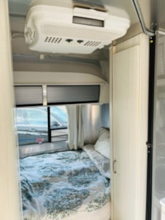 Inside this sleek RV rental: bedroom area with bed, windows, & overhead AC unit.