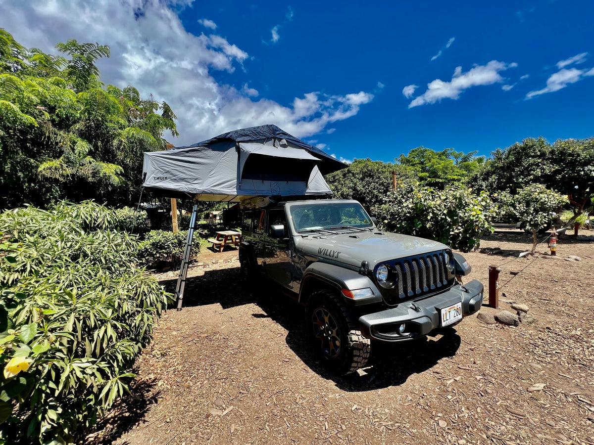 Gray Jeep with rooftop tent deployed at a sunny campsite. A cool RV rental for immersive outdoor adventures.
