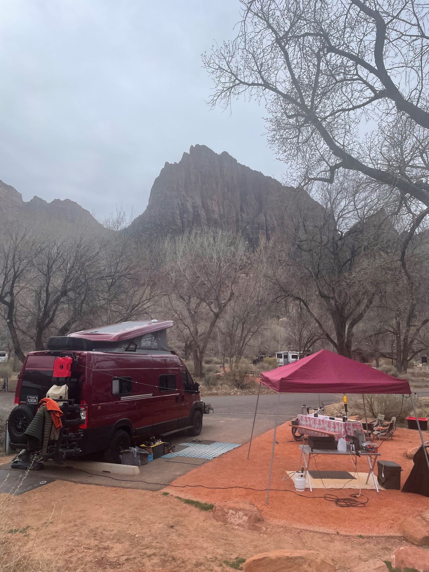 Scenic RV rental campsite with a red camper van, pop-up canopy, & outdoor kitchen setup amidst mountains and trees. Overcast day.