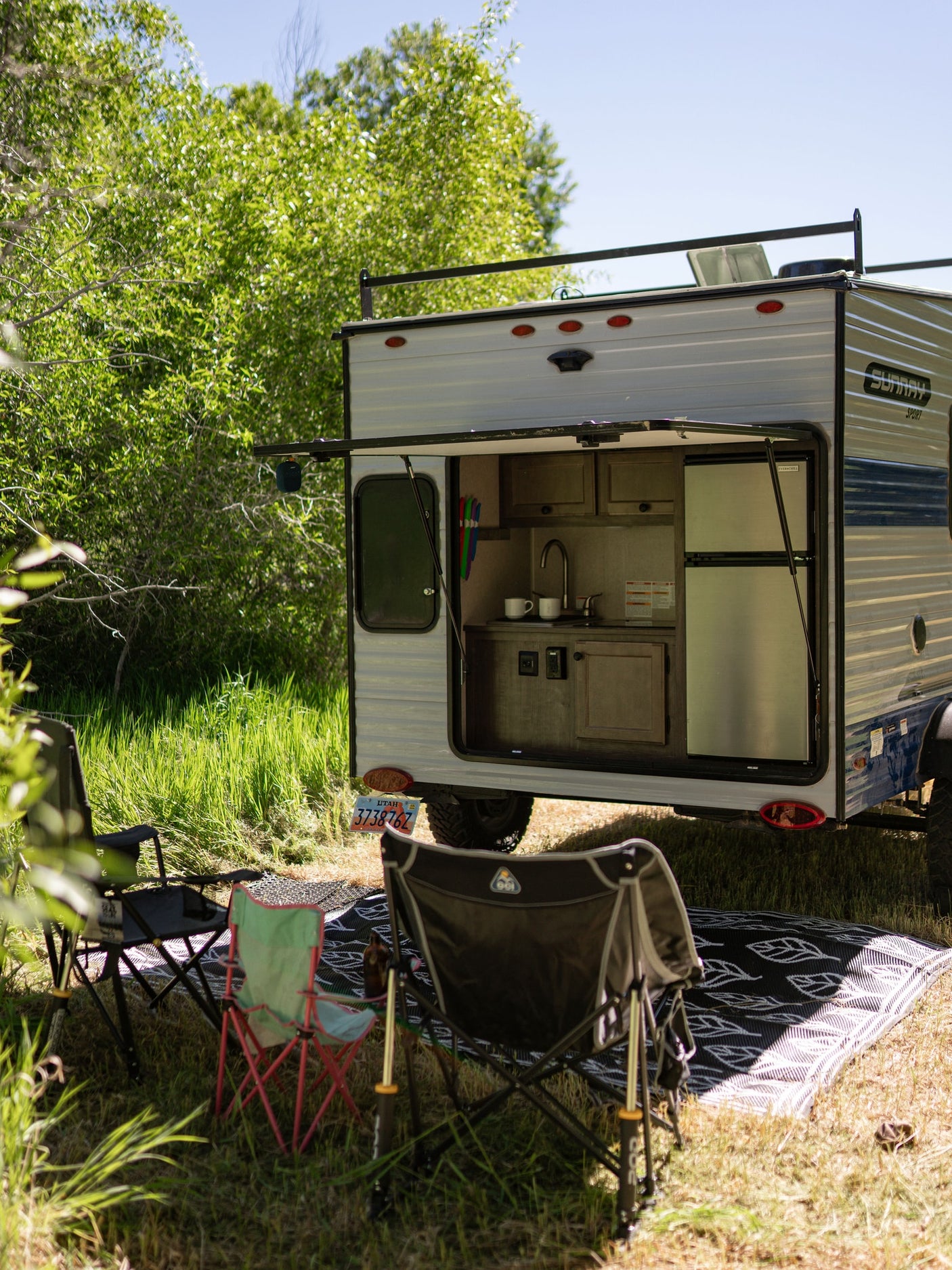 RV rental: Small camper with an open outdoor kitchen, chairs, and mat, set up at a sunny campsite amidst green foliage.