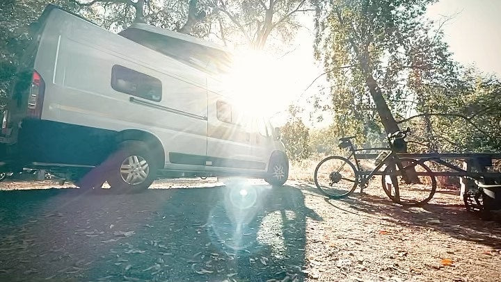 White RV rental camper van with pop-top, bike, and trailer, parked in a sun-drenched natural setting for adventure.