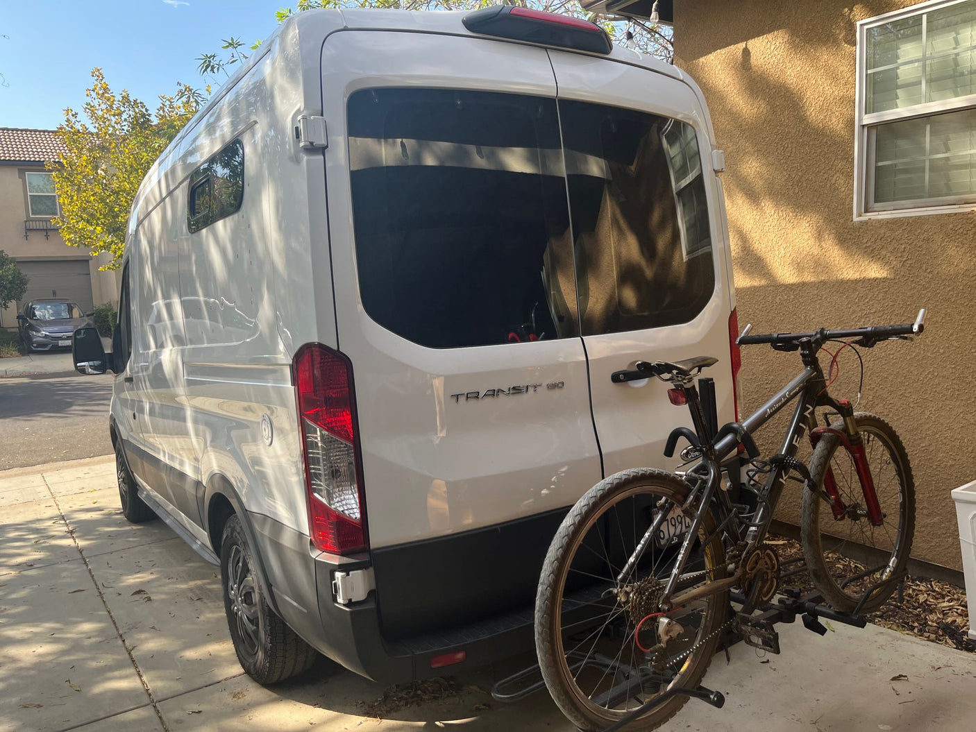 White Ford Transit camper van, equipped with a bike rack holding two mountain bikes, parked on a driveway. Ideal rv rental.