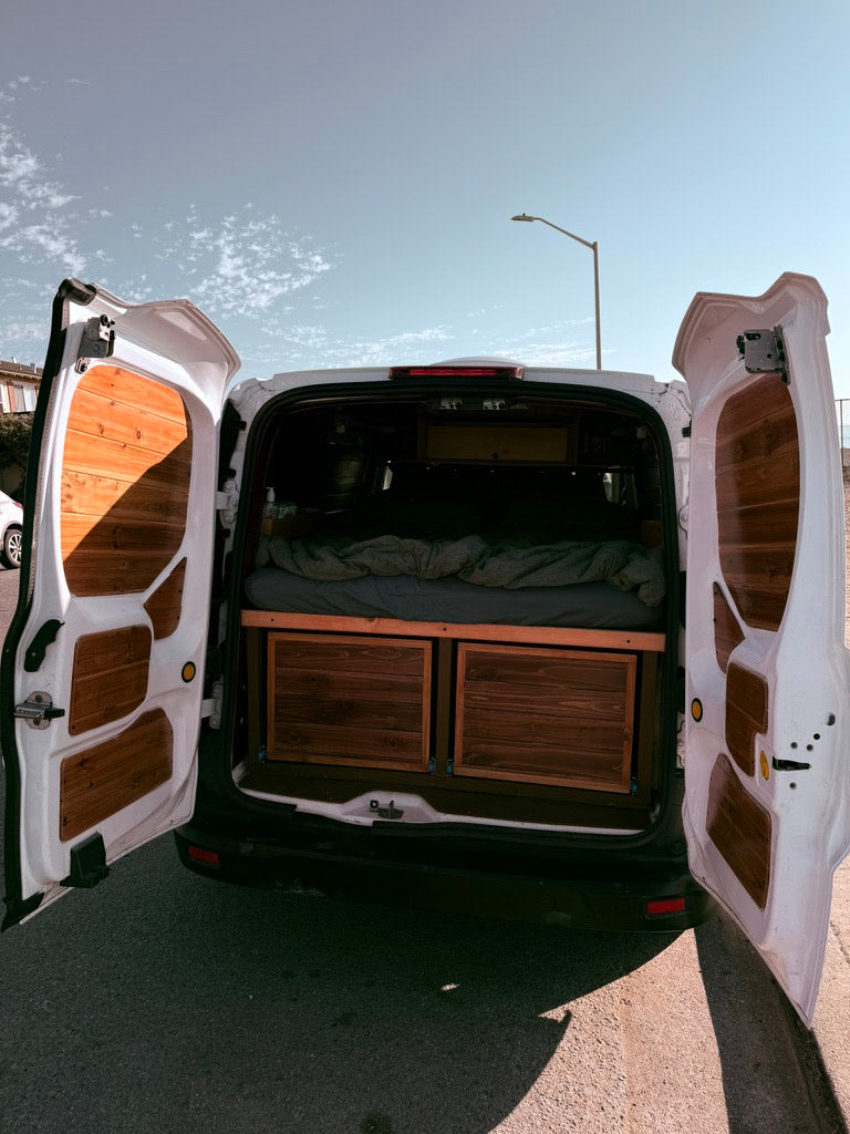 RV rental van: Open rear doors showcase a cozy bed with integrated wood storage, perfect for your next adventure.
