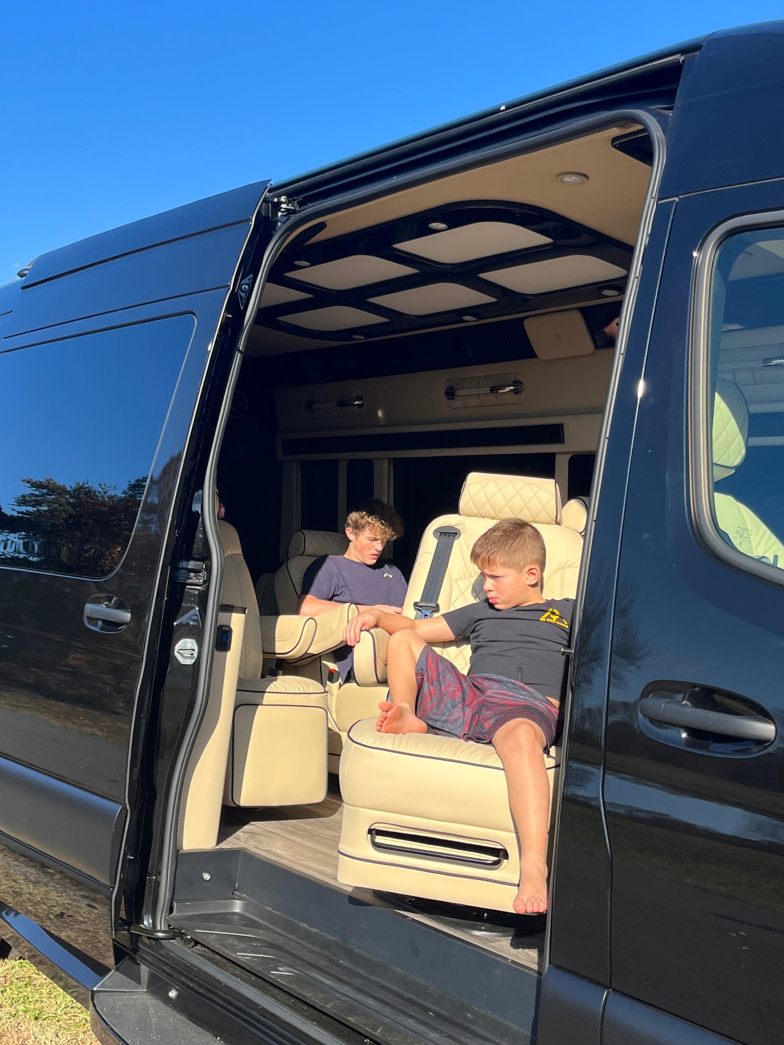 Two boys relax inside a premium RV rental van, showcasing its plush seating and custom interior via an open side door.