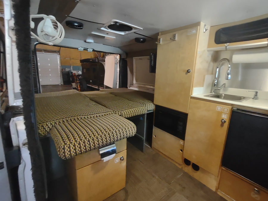View inside an RV rental, showing a multi-section bed/seating area, kitchen with sink, and light wood cabinetry.