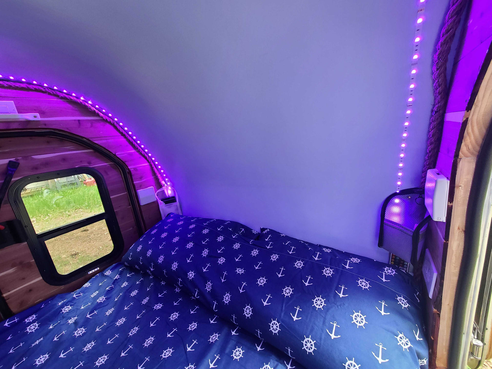Inside an RV rental bedroom, a bed with blue anchor sheets is illuminated by purple LED strip lights.