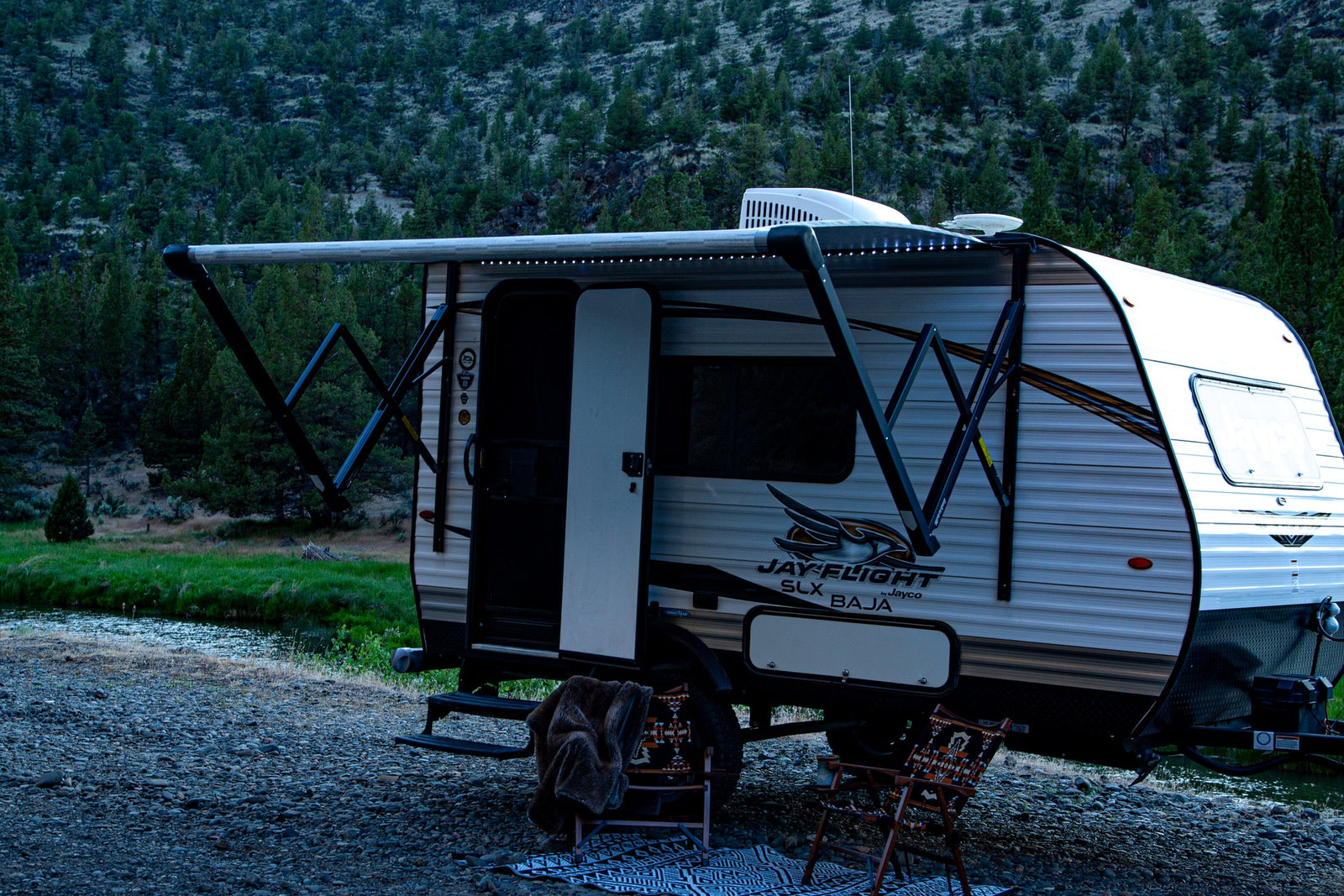 and two happy dogs at a beautiful outdoor campsite.|A truck towing a light travel trailer (Jayco Jay Flight SLX Baja) in front of an American flag mural. Perfect for your next rv rental trip!|An RV rental Jayco trailer and black truck parked roadside with an extended awning by a river & wooded mountain.|Riverside RV rental campsite at dusk
