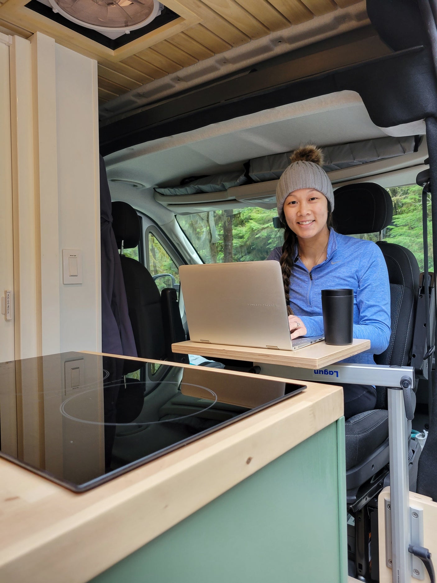 Smiling woman remote working in a cozy RV rental with a laptop, cooktop, & forest views.