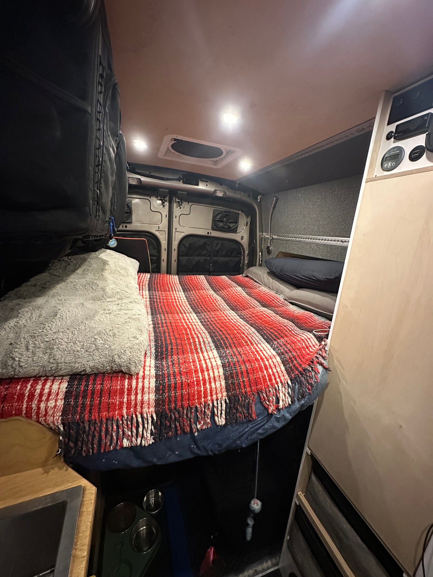 Well-lit rv rental camper van interior: a bed with a red plaid blanket, ceiling vent, and control panel.