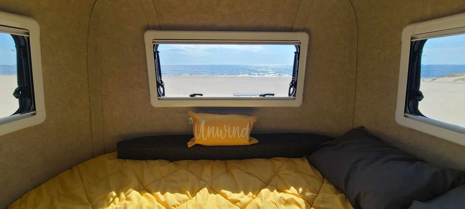 Serene rv rental interior: cozy bed with yellow comforter & "Unwind" pillow, offering a stunning beach and ocean view.