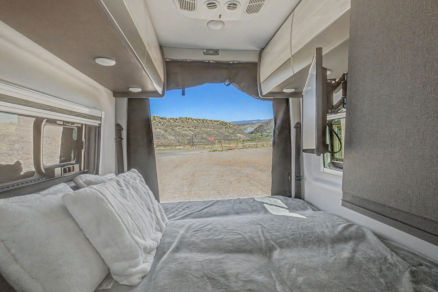 This rv rental features a cozy bed with soft pillows, offering a scenic view of hills and road from the open rear.