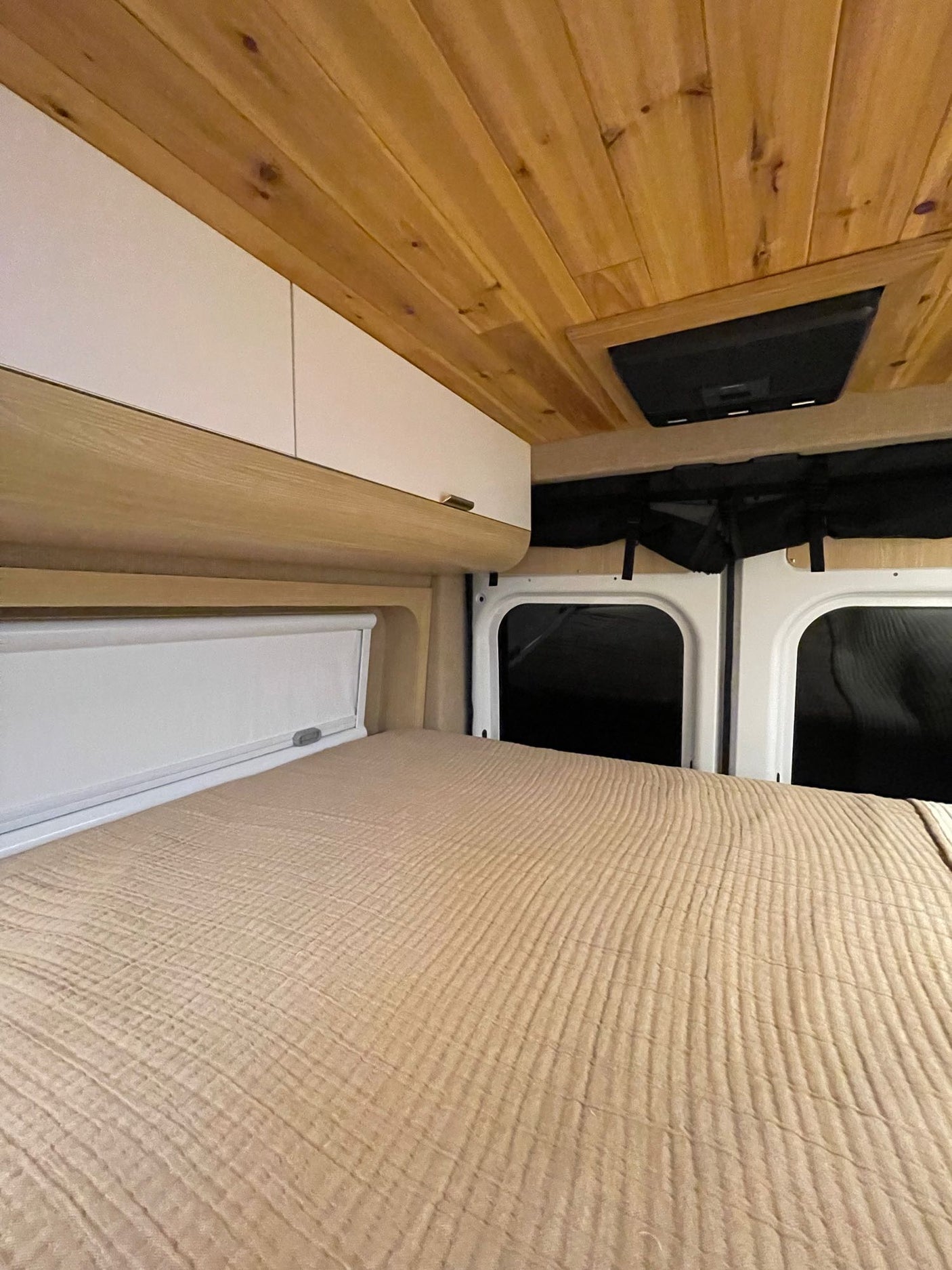Inside an RV rental: a beige bed, white overhead cabinets, wood ceiling, and black window coverings.