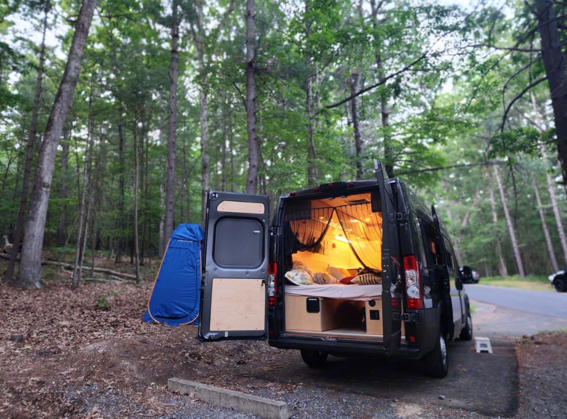 Cozy camper van RV rental set up for camping in a forest. Rear doors open reveal a bed and a blue privacy tent nearby.