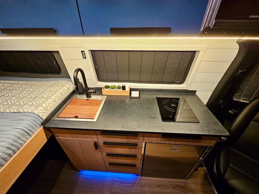 RV rental interior: a compact kitchen with a sink, induction cooktop, and fridge, beside a bed, all under LED lighting.
