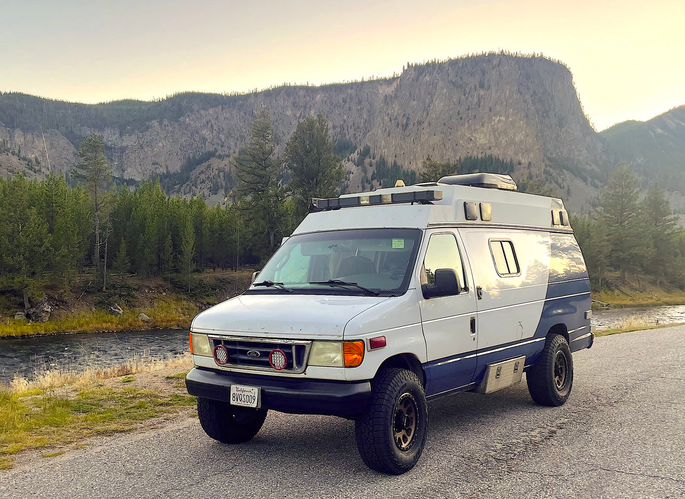 A white and blue custom Ford camper van, ideal for an RV rental, parked on a road next to a river with forested mountains.