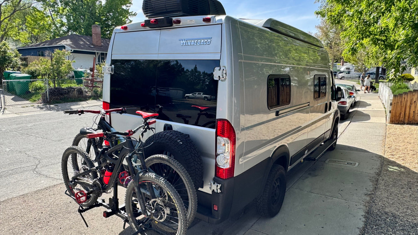 Winnebago RV rental with mountain bikes & spare tire, parked on a sunny street. Ideal for outdoor adventure travel.