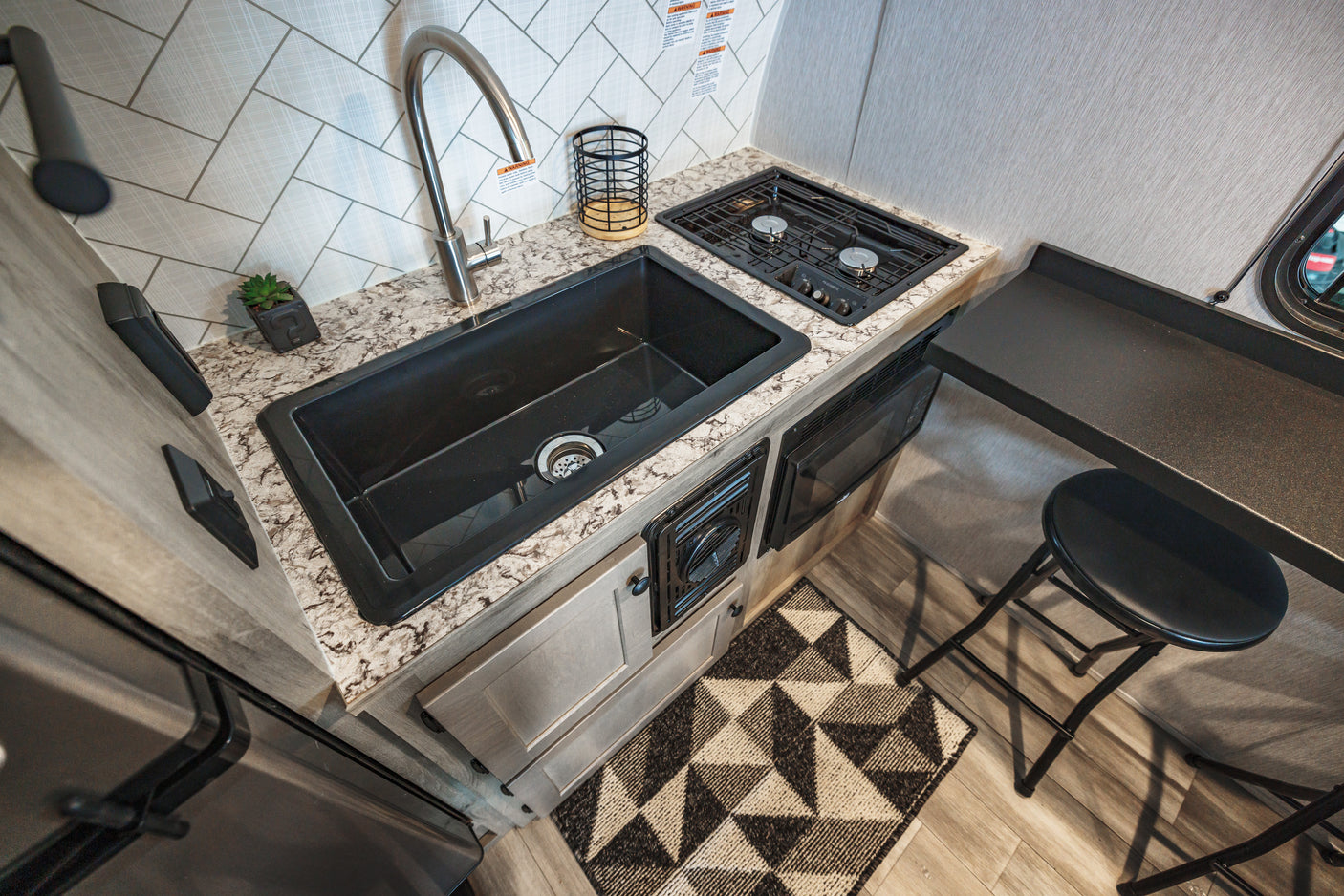 Modern RV rental kitchen features speckled counters, tiled backsplash, a dark sink, two-burner stove, microwave, and folding stool.