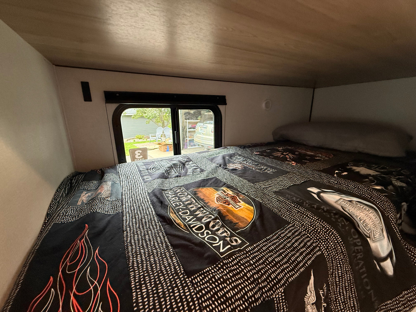 Cozy RV rental bed with a Harley-Davidson quilt, offering a window view of a residential area.