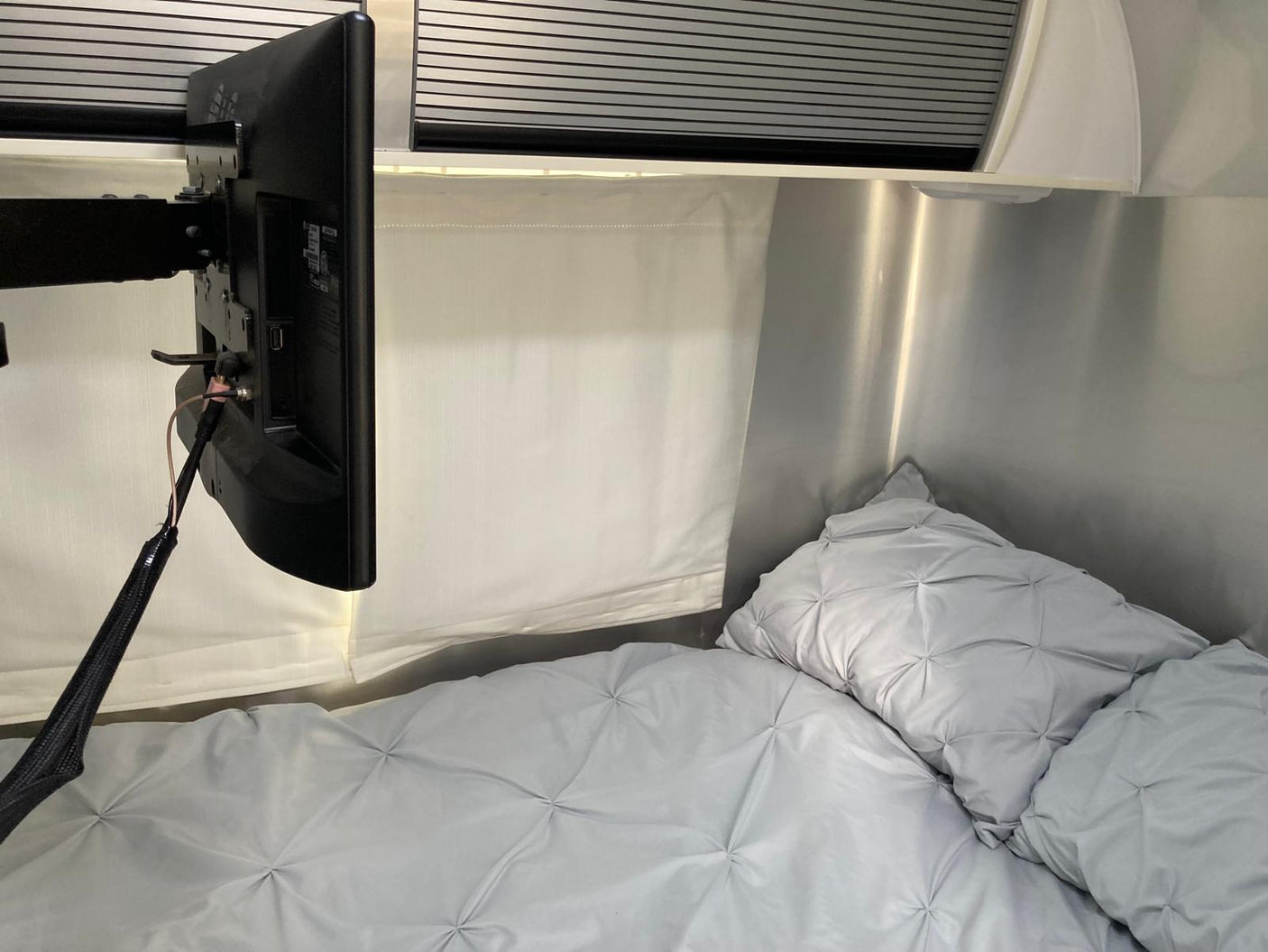 Cozy RV rental bedroom with a mounted TV, soft grey quilted bedding, and metallic walls. Ideal for comfortable travel.