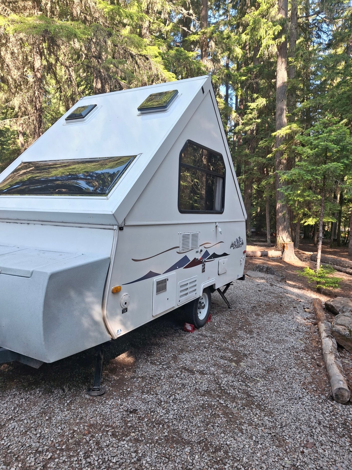 surrounded by tall trees.|White A-frame RV rental parked on a gravel campsite amidst tall forest trees.|White Chalet pop-up camper