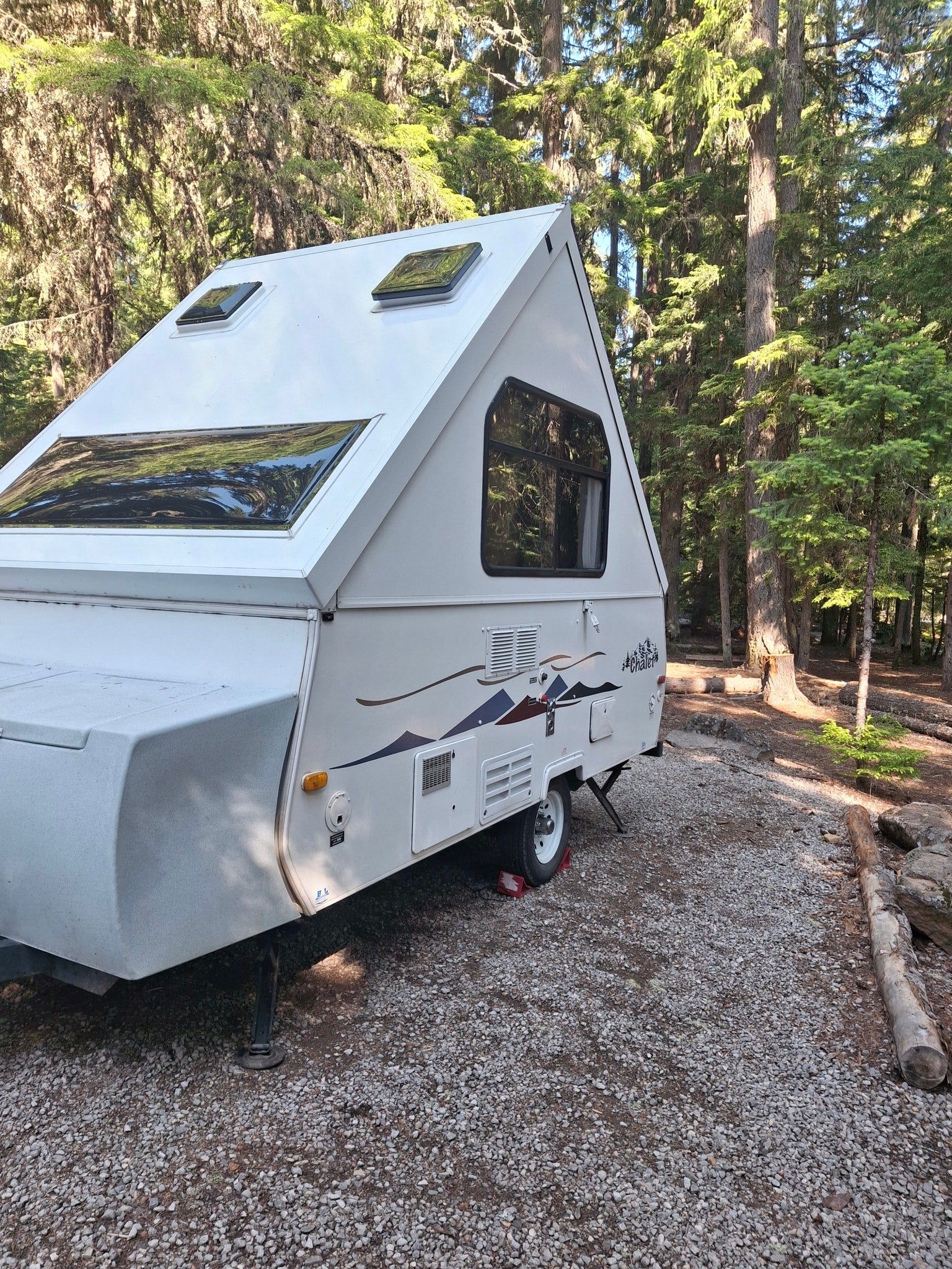 surrounded by tall trees.|White A-frame RV rental parked on a gravel campsite amidst tall forest trees.|White Chalet pop-up camper