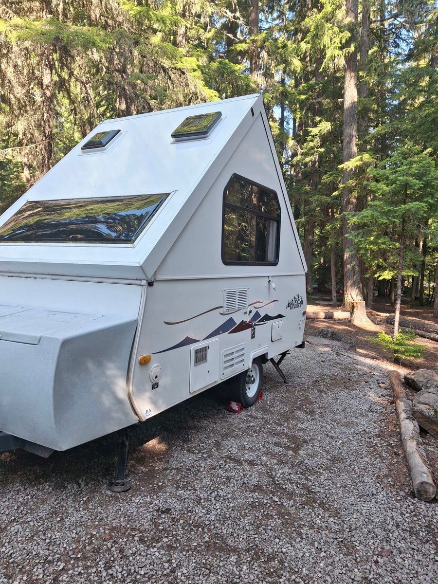 surrounded by tall trees.|White A-frame RV rental parked on a gravel campsite amidst tall forest trees.|White Chalet pop-up camper