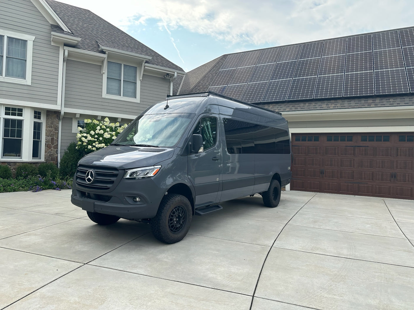 Modern grey Mercedes-Benz Sprinter van, ready for rv rental, parked on a concrete driveway in front of a house with solar panels.