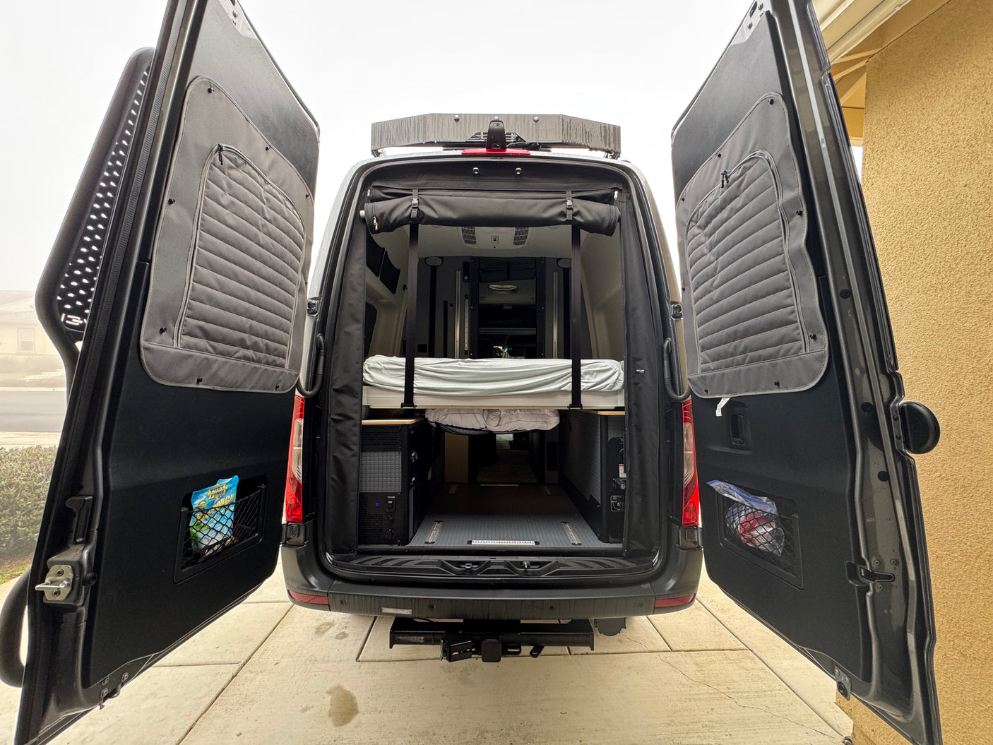 Rear view of an RV rental camper van with open doors, showcasing the interior bed, power system, and rear storage.