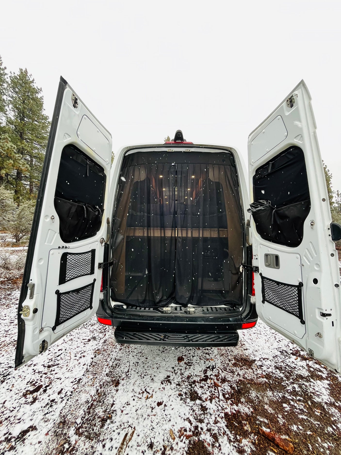 This RV rental van, with its rear doors open and a black mesh screen inside, is parked in a snowy forest setting.