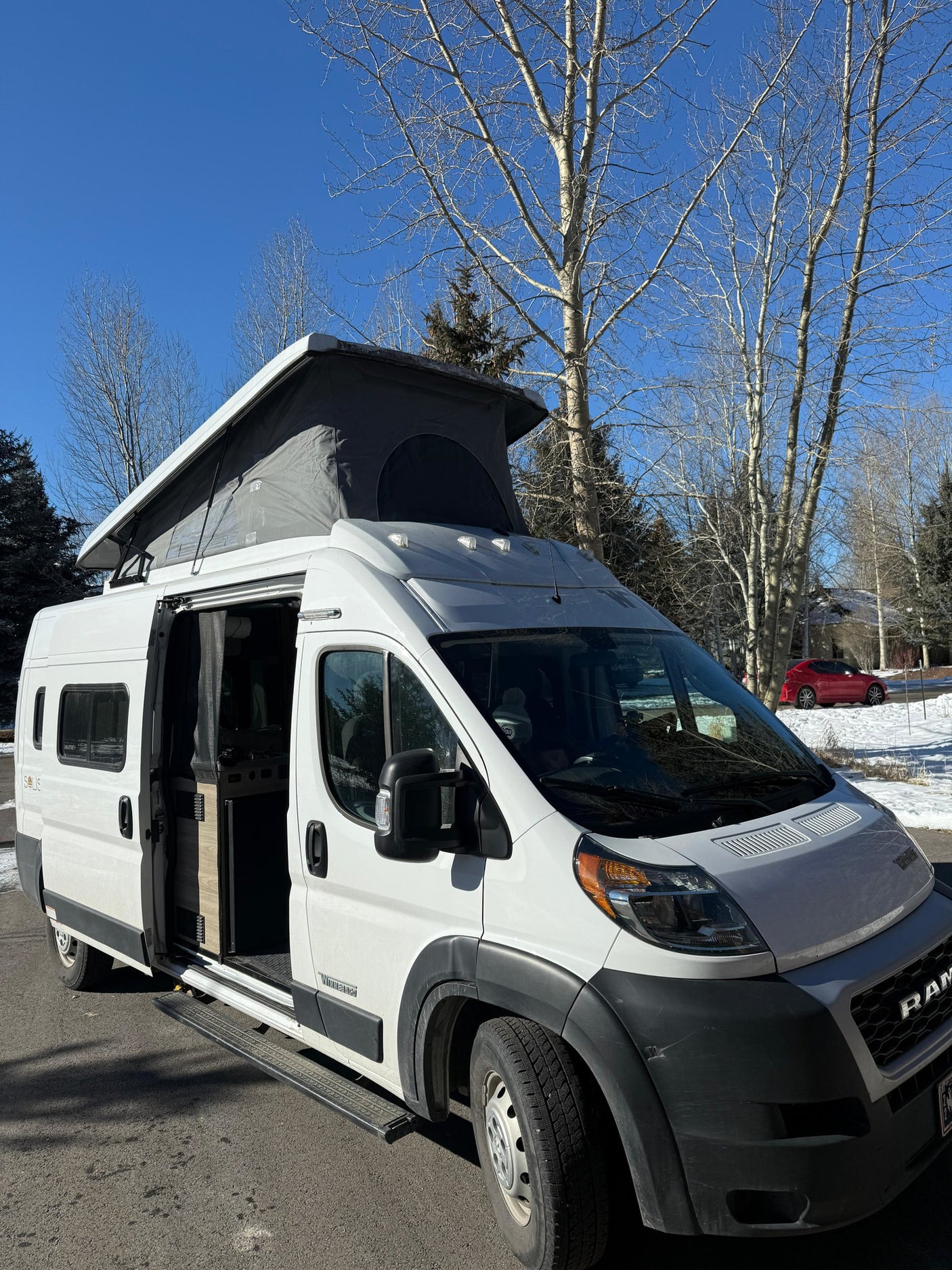 White pop-top camper van with open side door, ready for your rv rental. Sunny winter day, bare trees in background.