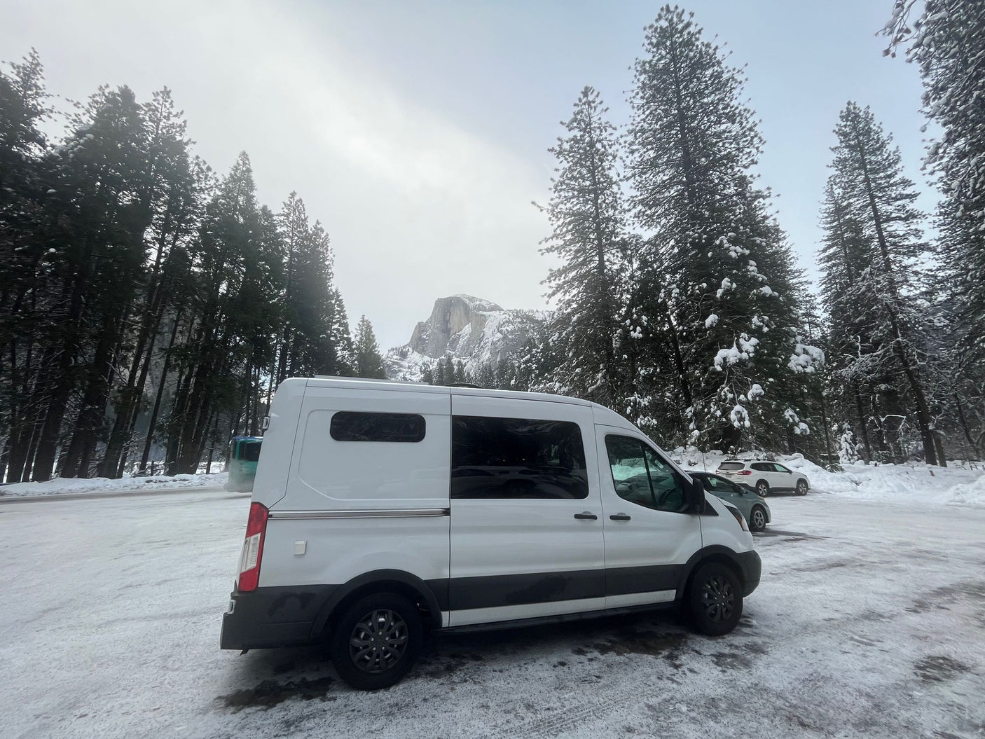 A white camper van (RV rental) is parked in a snowy lot, surrounded by tall pines, with Half Dome visible in the misty background.