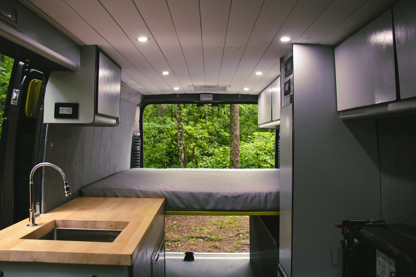 RV rental interior: kitchen with wooden counter, sink, faucet, bed, and forest view from the back.