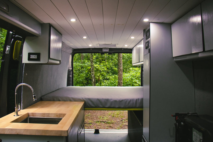 RV rental interior: kitchen with wooden counter, sink, faucet, bed, and forest view from the back.