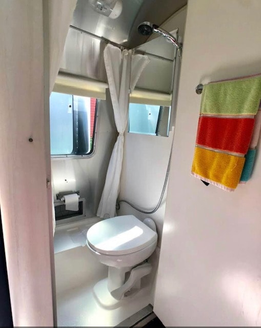 Inside an RV rental: a bright bathroom with toilet, shower, window, curtain, and striped towels hanging.