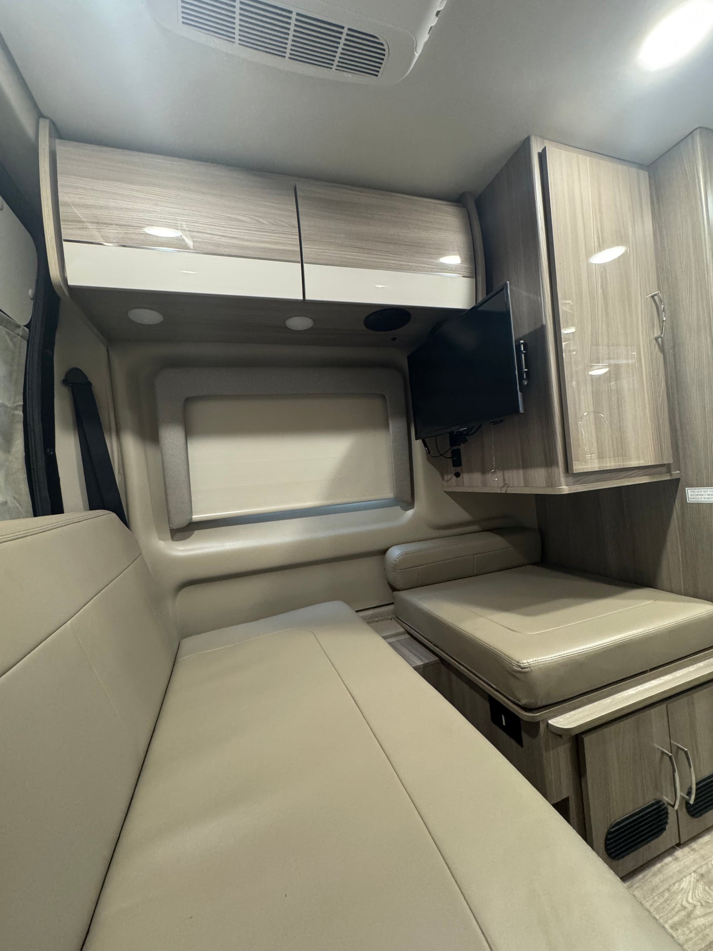 Step inside this RV rental featuring comfortable beige benches, wood-grain overhead cabinets, and a wall-mounted TV for your trip.