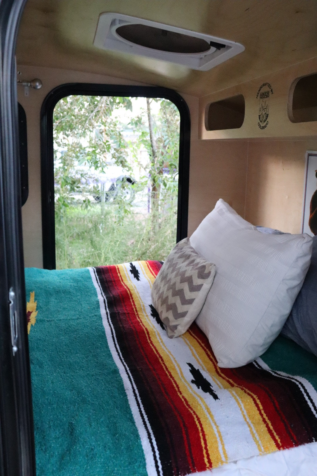 RV rental interior: cozy bed with colorful blanket and window view in a handcrafted Hiker Trailer.