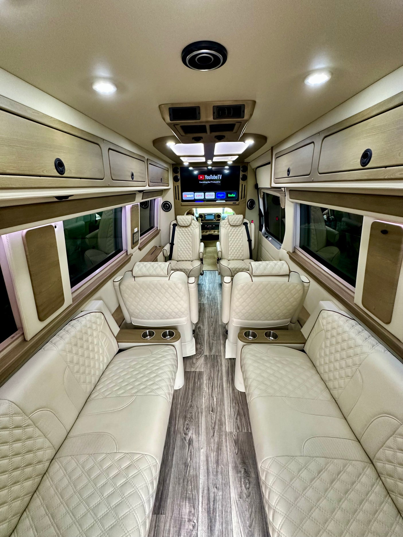 Luxury RV rental interior featuring cream quilted leather seats, wood-look flooring, overhead storage, and a large entertainment screen.