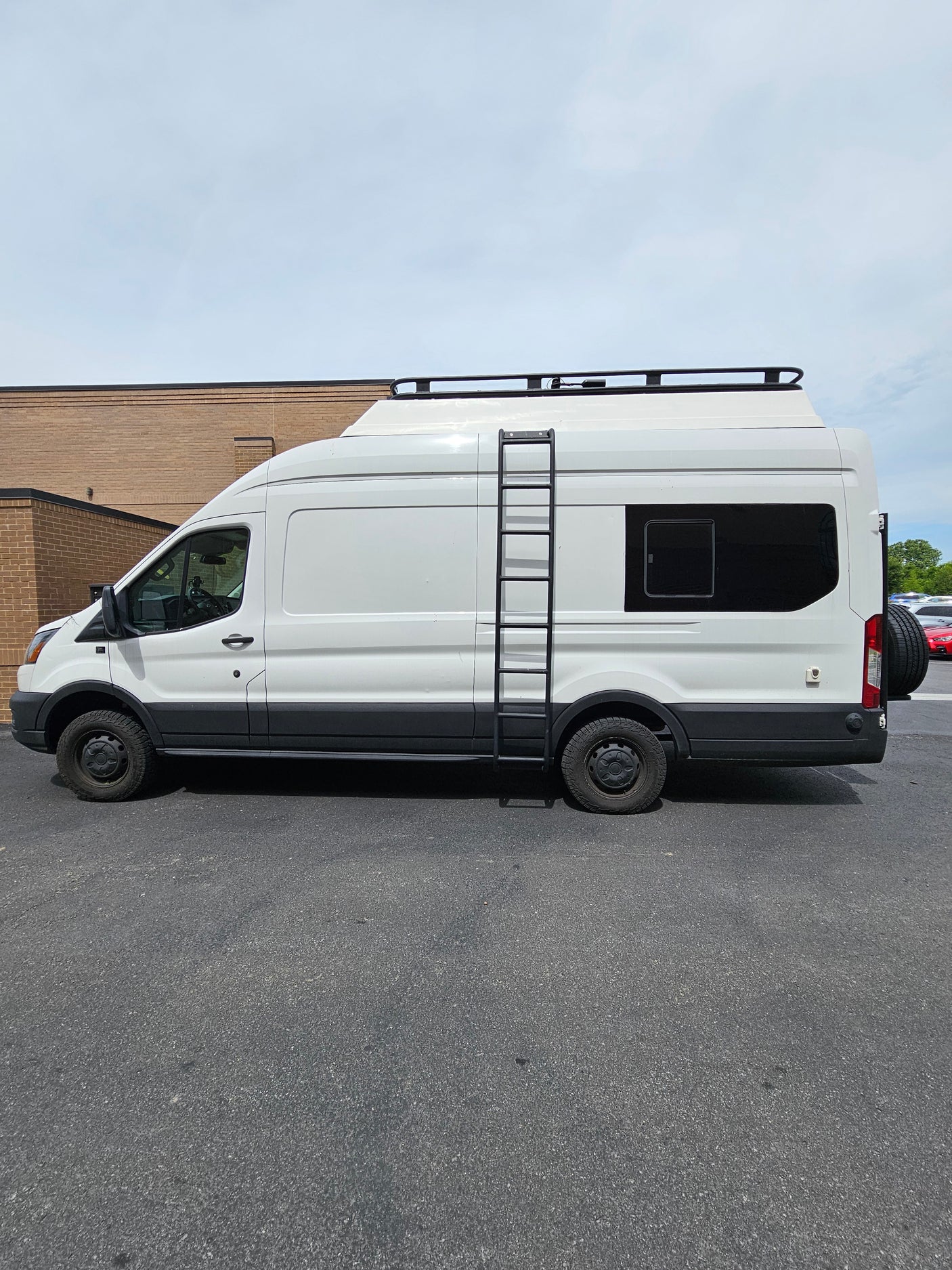 White Ford Transit RV rental camper van, featuring a roof rack, side ladder, and spare tire on the back.
