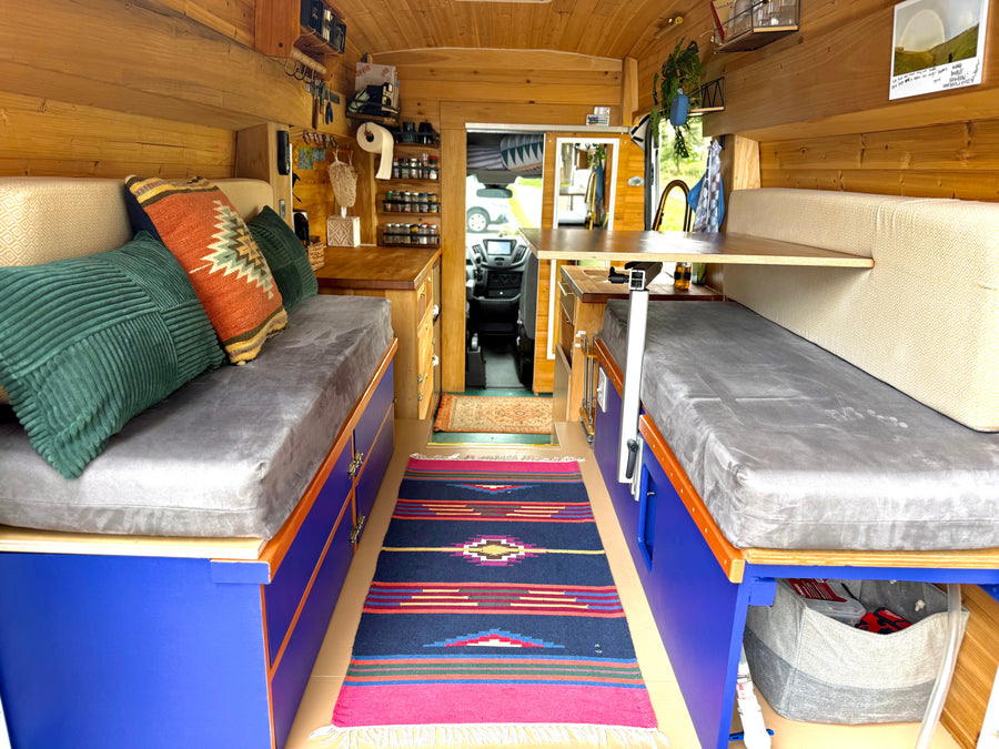 Cozy RV rental interior: wood-lined, vibrant blue storage, grey cushions, colorful pillows, and a compact kitchen. Road trip ready!