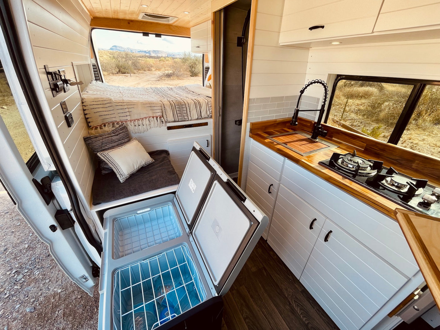 RV rental: Bright camper van interior with bed, kitchen, and open dual-zone portable fridge, parked in desert.