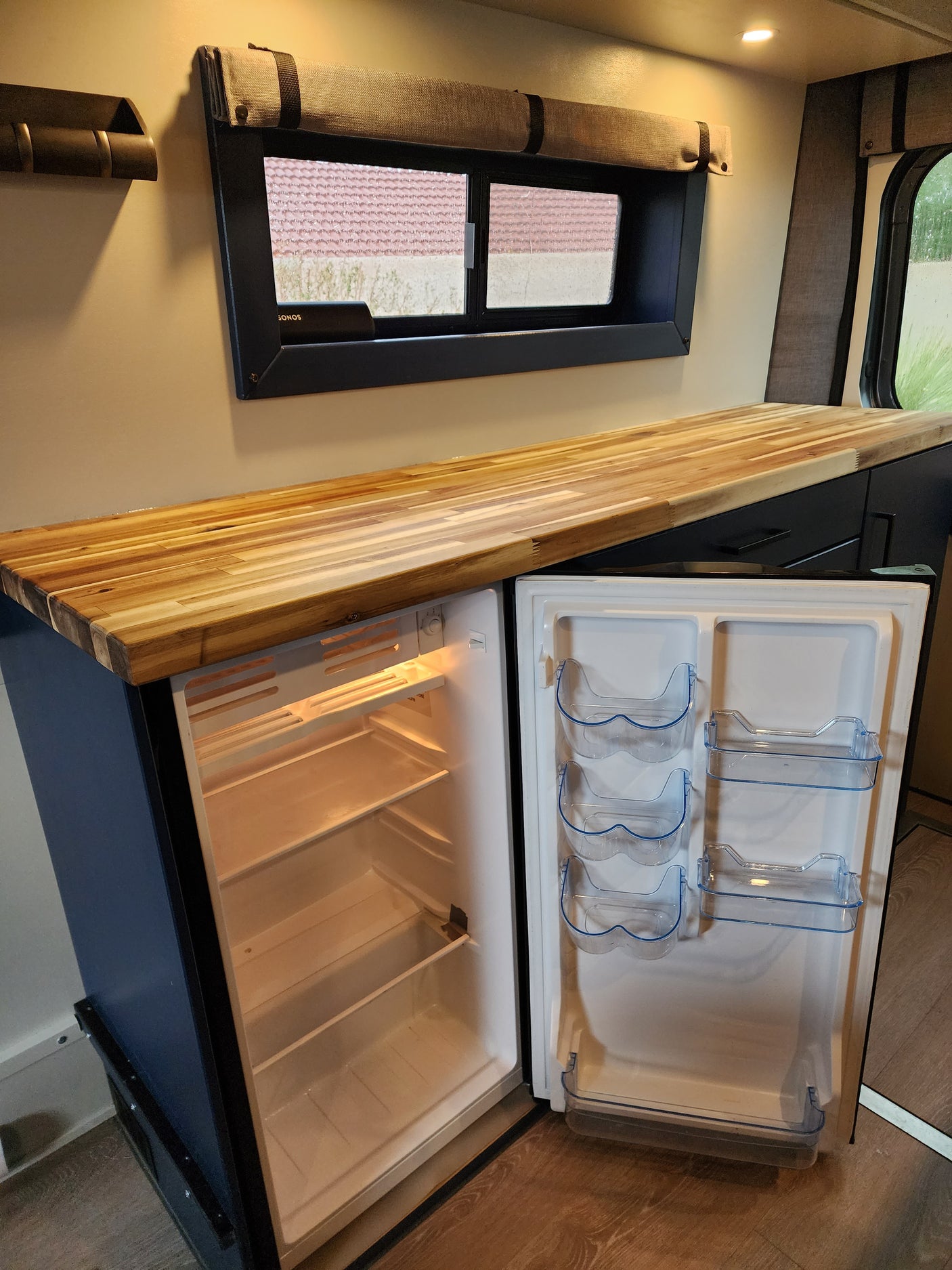 See a bright, clean RV rental interior with an open mini-fridge, wooden counter, and window with a rolled-up shade.