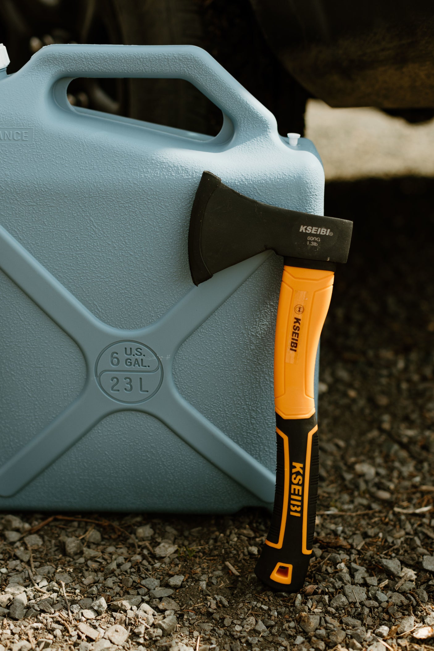 A light blue 6-gallon water jug and an orange-handled KSEIBI axe rest on gravel, ready for your RV rental adventure.