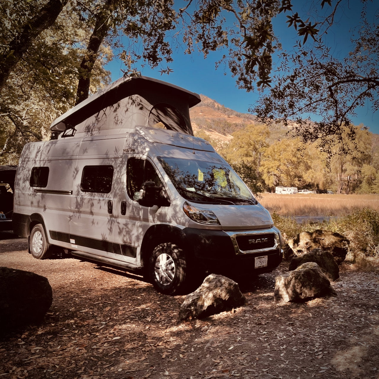 Experience outdoor camping in this gray rv rental camper van with a pop-top. Parked among trees, with mountains & water in view.