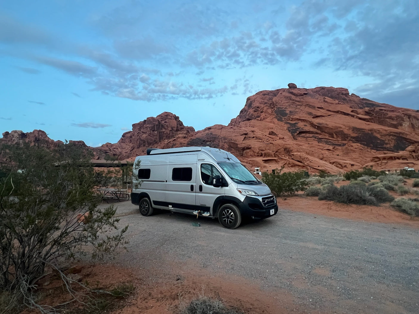 A silver camper van, possibly an RV rental, parked on a dirt road amidst stunning red rock formations and desert plants.