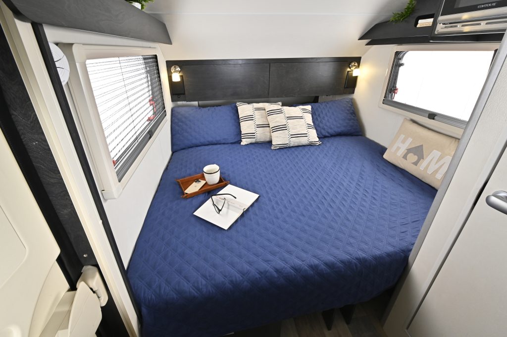 Cozy RV rental bedroom with a large bed, blue quilted bedding, two windows, and a reading setup for comfortable journeys.