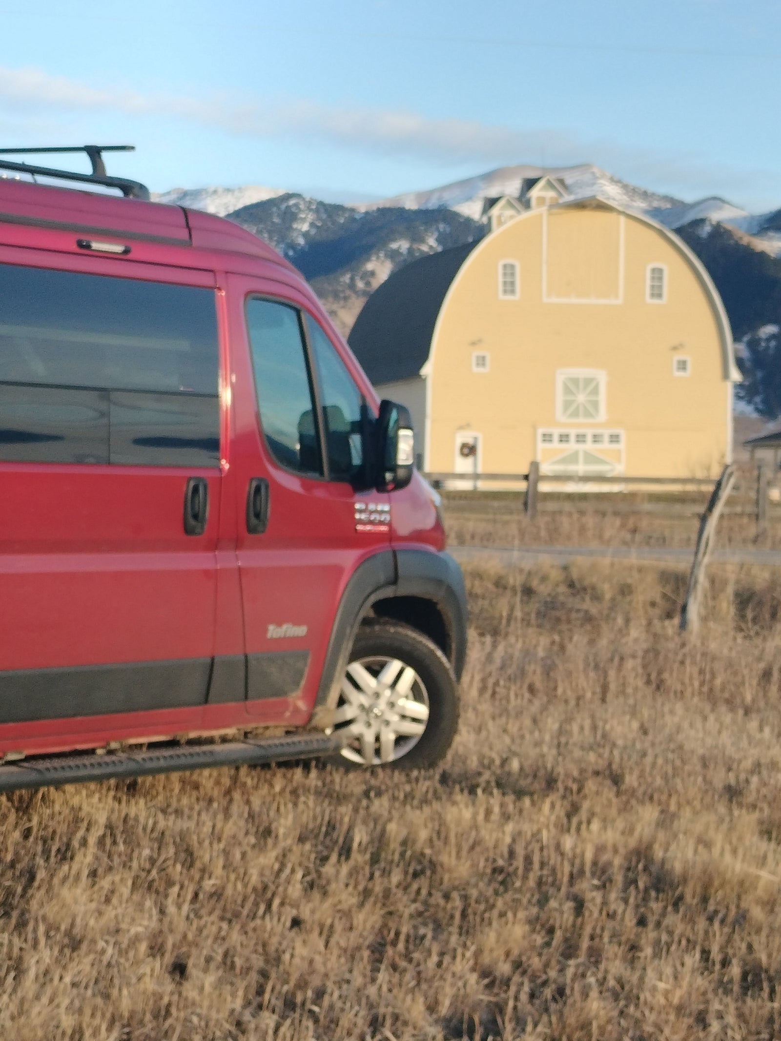 Red RV rental van in a dry field, with a large yellow barn and snowy mountains.